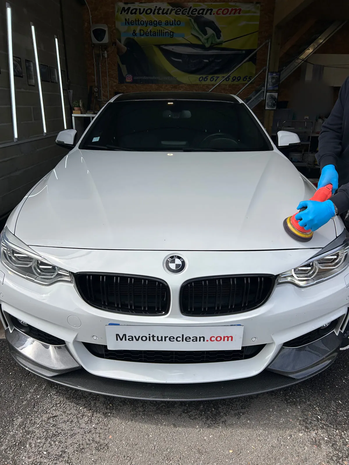 Polisseuse orbitale tenue par des mains gantées de bleu sur le capot d'une BMW blanche, à l'entrée de l'atelier